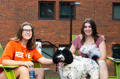 Two students sitting outside in green chairs with a black and white dog